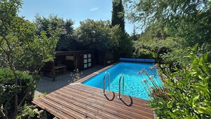 Eine Holzterrasse mit Swimmingpool, umgeben von Grünpflanzen und einem kleinen Gebäude mit einem Fenster.