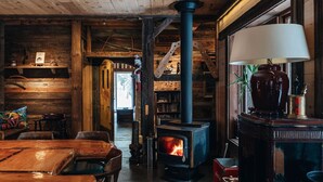 A rustic wooden interior with a central wood-burning stove, a dining area with wooden tables and chairs, and a bookshelf in the background.