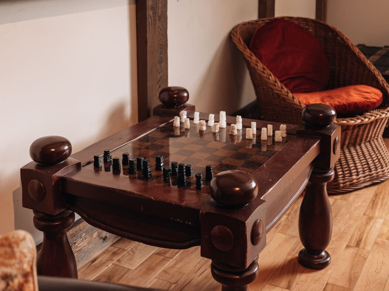 A wooden chess table with a checkered board and pieces, positioned in a room with wooden flooring and a wicker chair with a red cushion in the background.