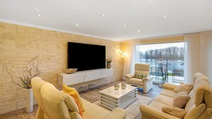 A modern living room with a flat-screen TV, beige sofas, a coffee table, and a large window with a view. - Apartment 'Suite Seeblick' with Lake View, Private Garden and Wi-Fi (Holzappel)