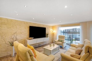 A modern living room with a flat-screen TV, beige sofas, a coffee table, and a large window with a view. - Apartment 'Suite Seeblick' with Lake View, Private Garden and Wi-Fi (Holzappel)