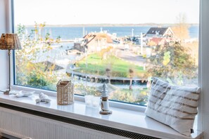 A window with a view of a coastal town, a lamp, a cushion, and a small decorative item on the sill. - KüstenGlück Langbaligau - Where the sea touches the heart. (Langballigau)