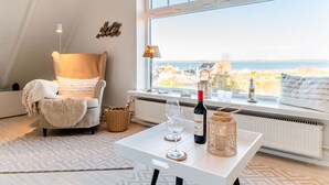 Ett mysigt rum med utsikt över havet, en fåtölj, ett bord med en flaska och glas, och en lampa.
