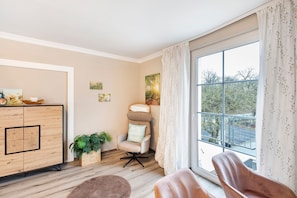 A room with a wooden cabinet, a cushioned chair, a window with a view of trees, and a plant.