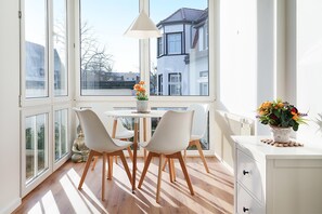 A bright, sunlit room with a dining table and chairs, a window with a view of buildings, and a white cabinet with drawers. - Cozy vacation apartment with Wi-Fi & parking—just 450 m from the sandy beach (Bansin)
