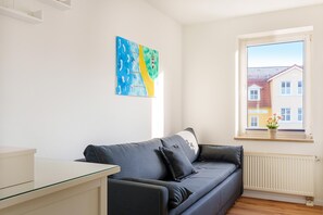 A modern living room with a sofa, a glass-top table, a radiator, and a window with a view of a building. - Cozy vacation apartment with Wi-Fi & parking—just 450 m from the sandy beach (Bansin)