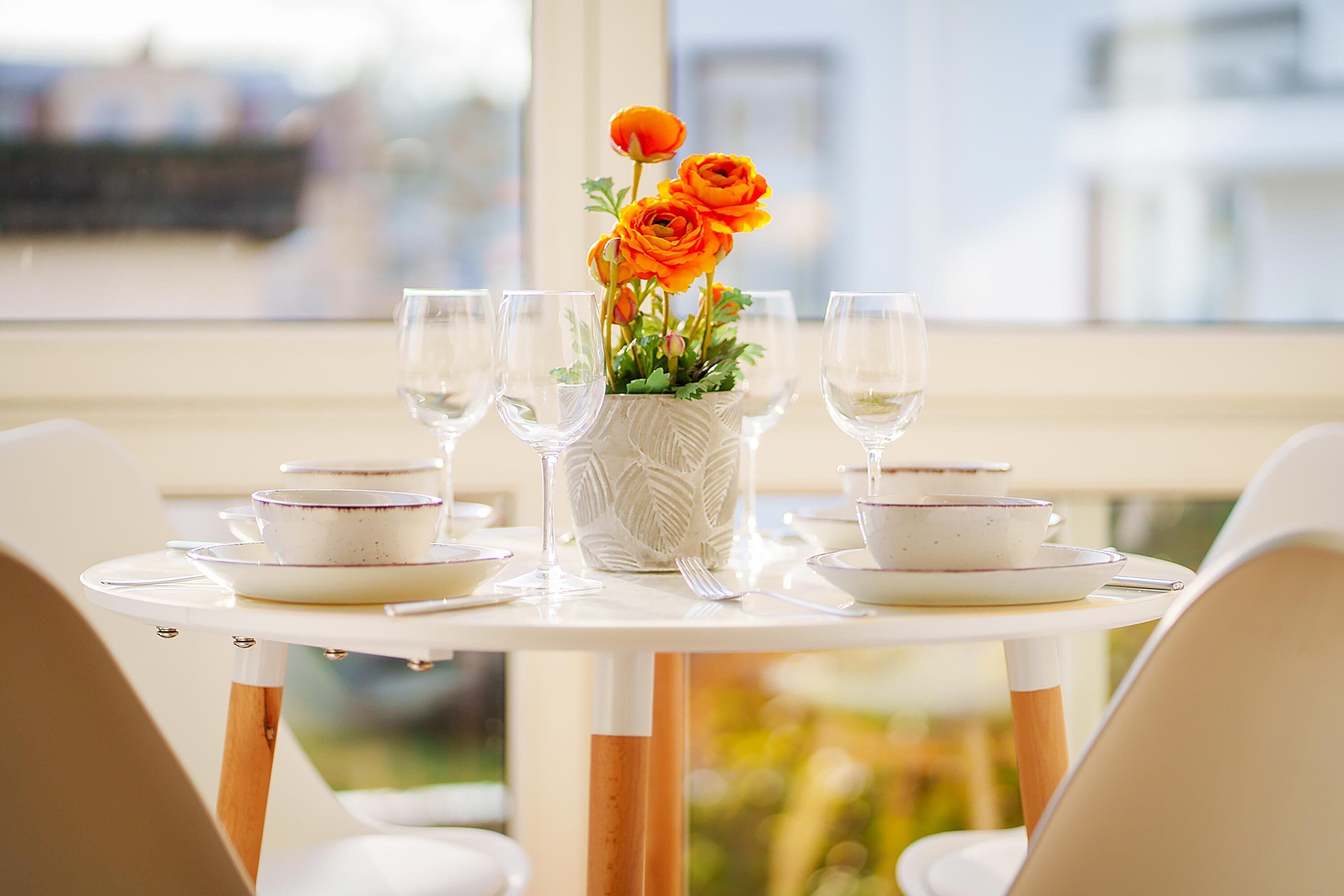 A dining table set with white plates, bowls, and glasses, centered with a vase of orange flowers.