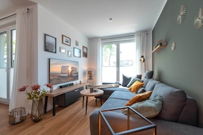 Un salon moderne avec un canapé, une table basse et un téléviseur. Des tableaux encadrés ornent le mur, et une fenêtre est agrémentée de rideaux.