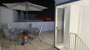 Een terras buiten met een tafel, stoelen, een barbecue en een parasol.