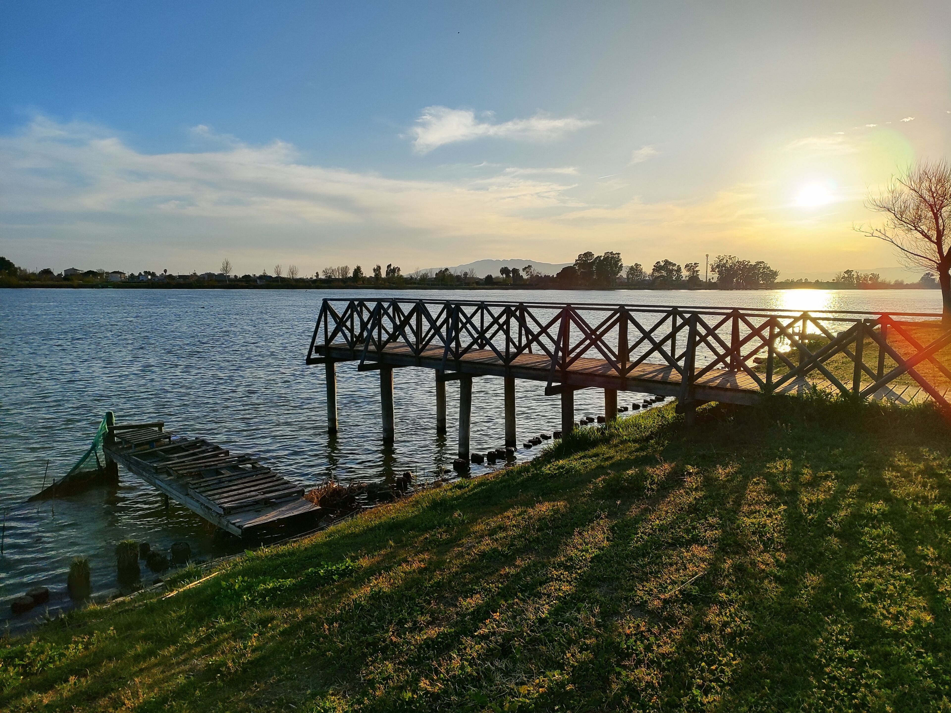 Une jetée en bois s’étendant au-dessus d’une étendue d’eau calme, avec une rive herbeuse d’un côté et un ciel dégagé au-dessus.