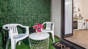 A room with a green artificial wall, a white round table with a flower vase, two white chairs, and a view into a kitchen area. - Apartment 'Il Tulipano House' with Private Terrace, Wi-Fi and Air Conditioning (Catania)