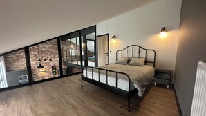 A bedroom with a loft-style design, featuring a bed, bedside tables, and a view of a dining area through large windows. - Au 171 Le loft avec un Sauna (Flémalle)