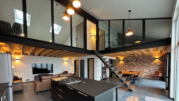 A spacious kitchen with a central island, a dining area with a brick wall, and a staircase connecting two levels. - Au 171 Le loft avec un Sauna (Flémalle)