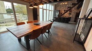 A spacious dining area with a large wooden table and modern chairs, a kitchen with a brick wall, and a staircase leading to an upper level. - Au 171 Le loft avec un Sauna (Flémalle)