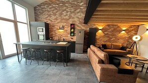 A modern kitchen with a large island, bar stools, and a built-in oven. There is a seating area with a sofa and a coffee table. - Au 171 Le loft avec un Sauna (Flémalle)