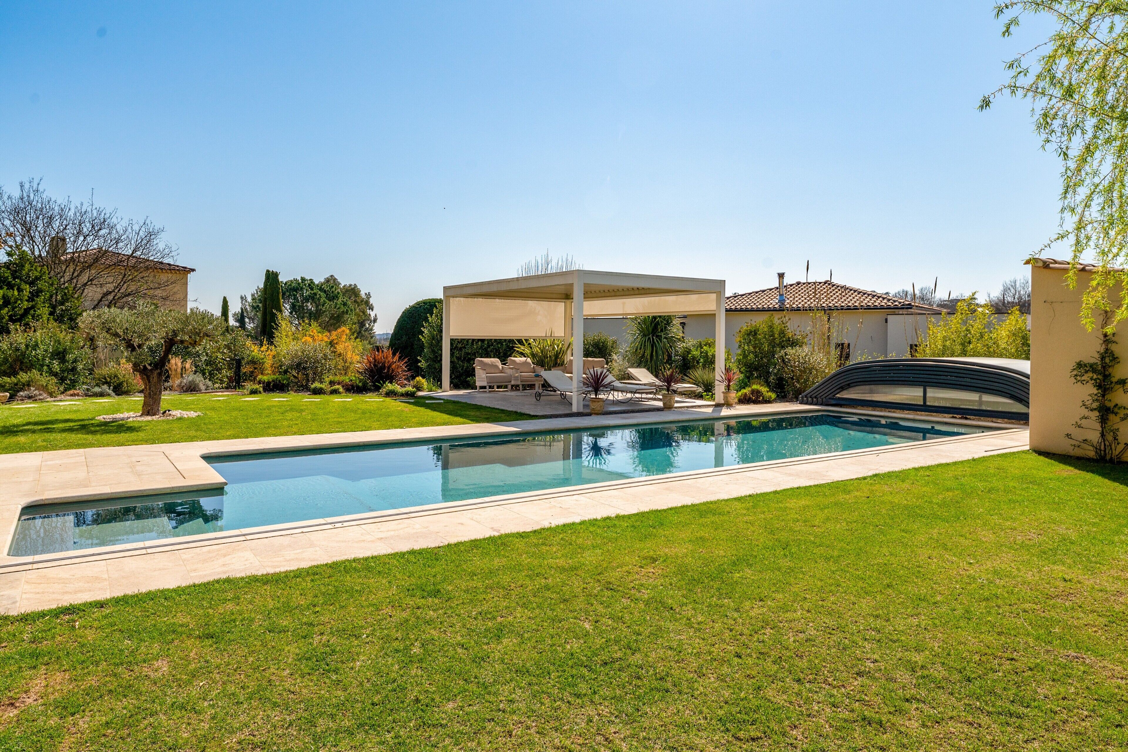 Una piscina con zona de estar cubierta, rodeada de un césped cuidado y diversas plantas.
