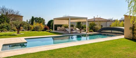Una piscina con zona de estar cubierta, rodeada de un césped cuidado y diversas plantas.
