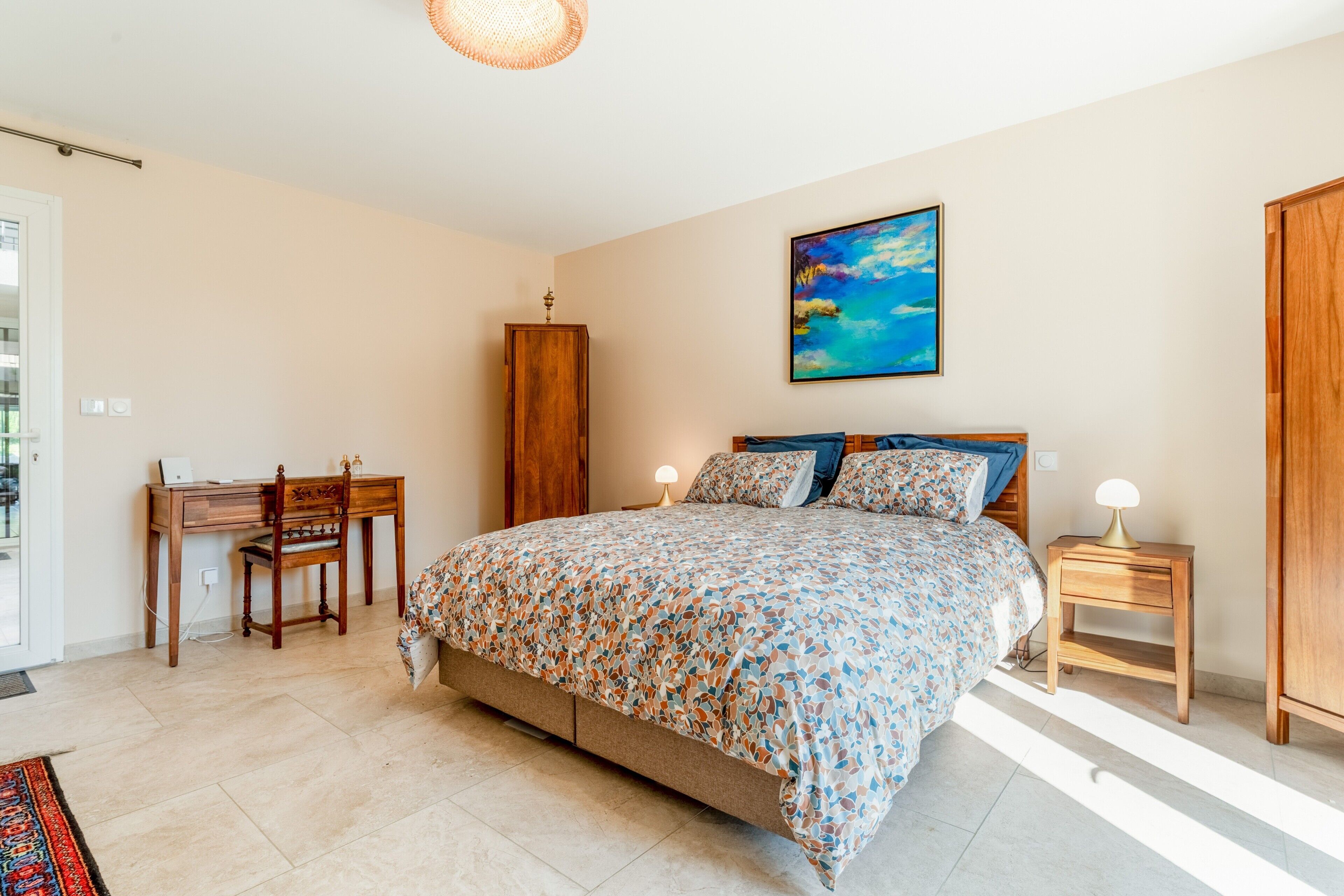 A bedroom with a bed, a desk, a chair, a nightstand, and a painting on the wall.