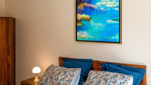 Un dormitorio con cabecera de madera, dos almohadas con estampado floral, una lámpara de noche y un cuadro abstracto y colorido en la pared.