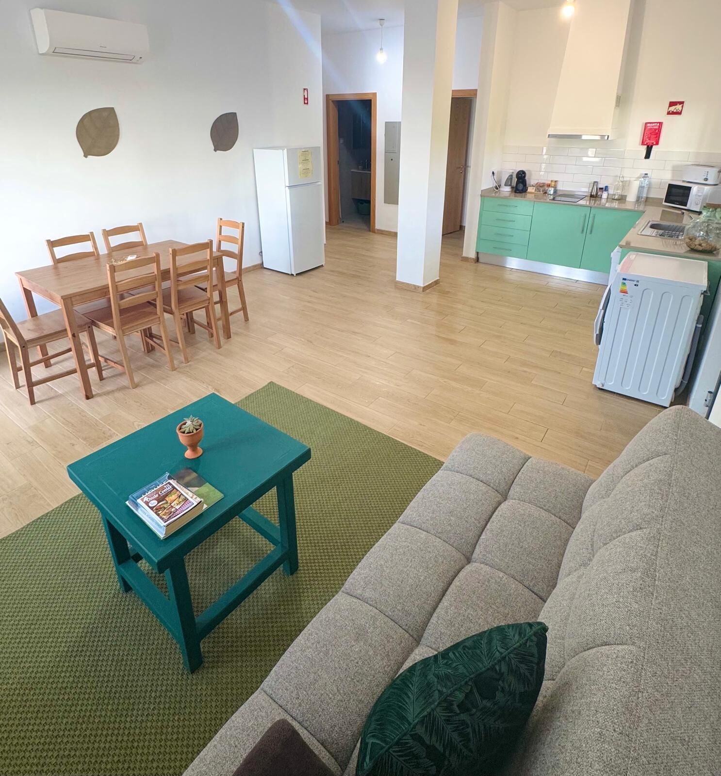 A living room with a sofa, a dining table with chairs, a kitchen area with a fridge, and a green rug.