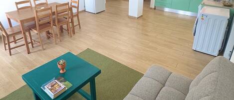 A living room with a sofa, a dining table with chairs, a kitchen area with a fridge, and a green rug.