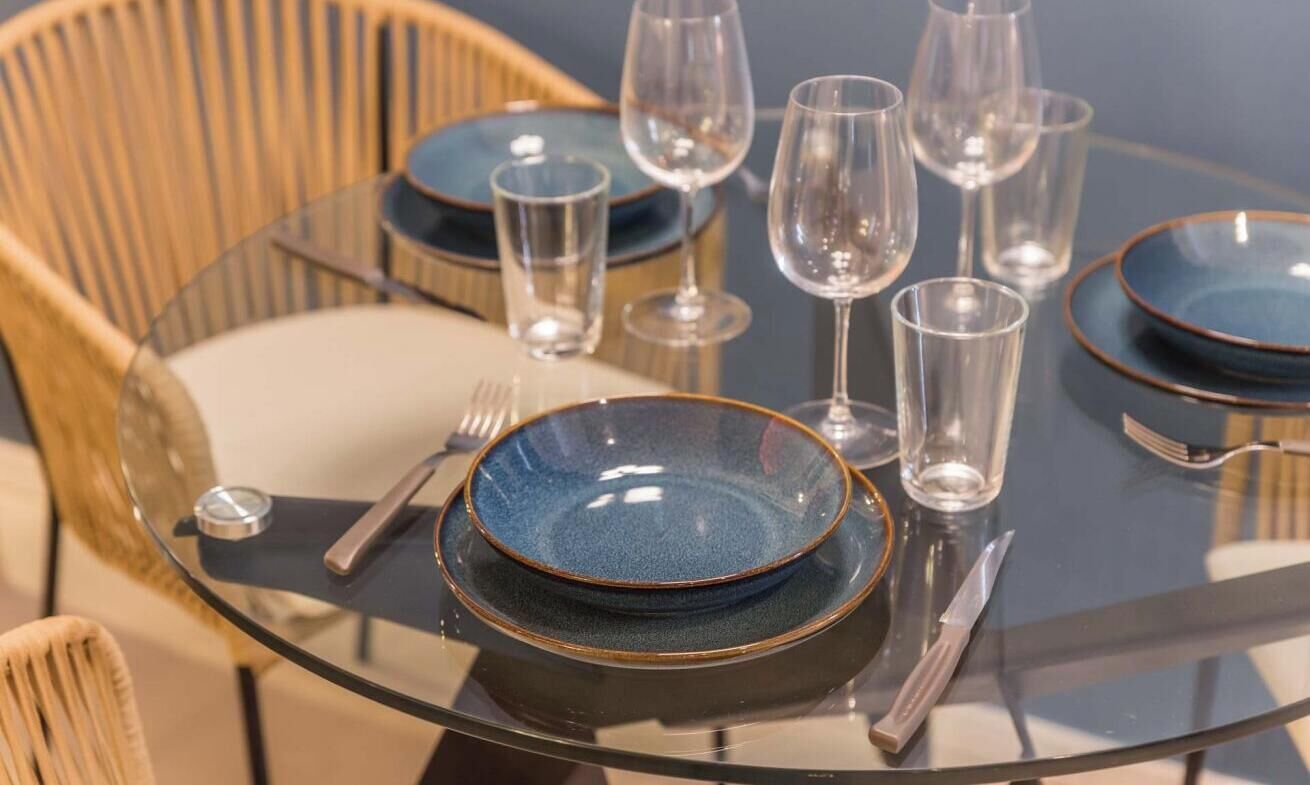 Una mesa de vidrio redonda con platos azules, vasos transparentes y cubiertos.