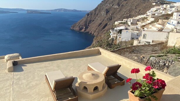 A terrace with two wooden lounge chairs and a small table, overlooking a coastal village with white buildings and a view of the sea. - Kamari Real Living, with Caldera views and Jacuzzi (Manolas)