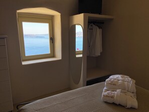 A room with a window overlooking a body of water, a bed with a textured white cover, a white towel with 'KAMARI' embroidered on it, and a white cabinet. - Kamari Real Living, with Caldera views and Jacuzzi (Manolas)