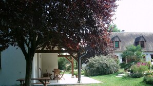 A covered outdoor seating area with tables and chairs, a tree, and a building with a sloped roof in the background. - Charming house with shared pool in Contres (Contres)