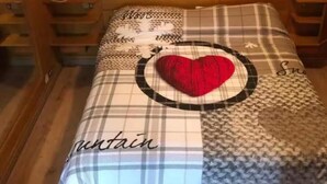 Un dormitorio con cabecera de madera, cama con colcha escocesa y almohada con forma de corazón, mesita de noche y un cuadro en la pared con un paisaje de montaña.