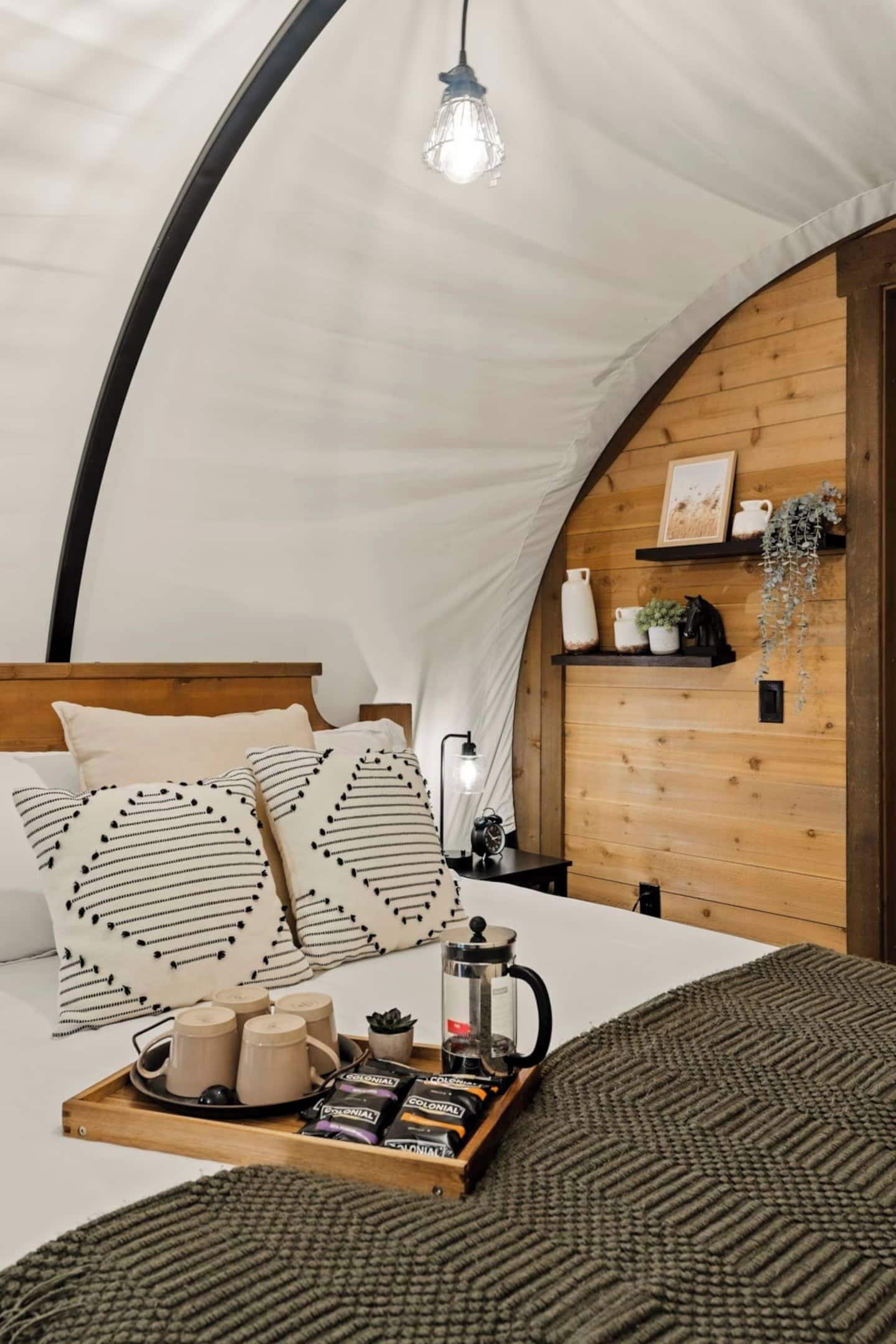 A cozy tent interior with a wooden bed, a tray of snacks, a coffee maker, and a hanging plant.