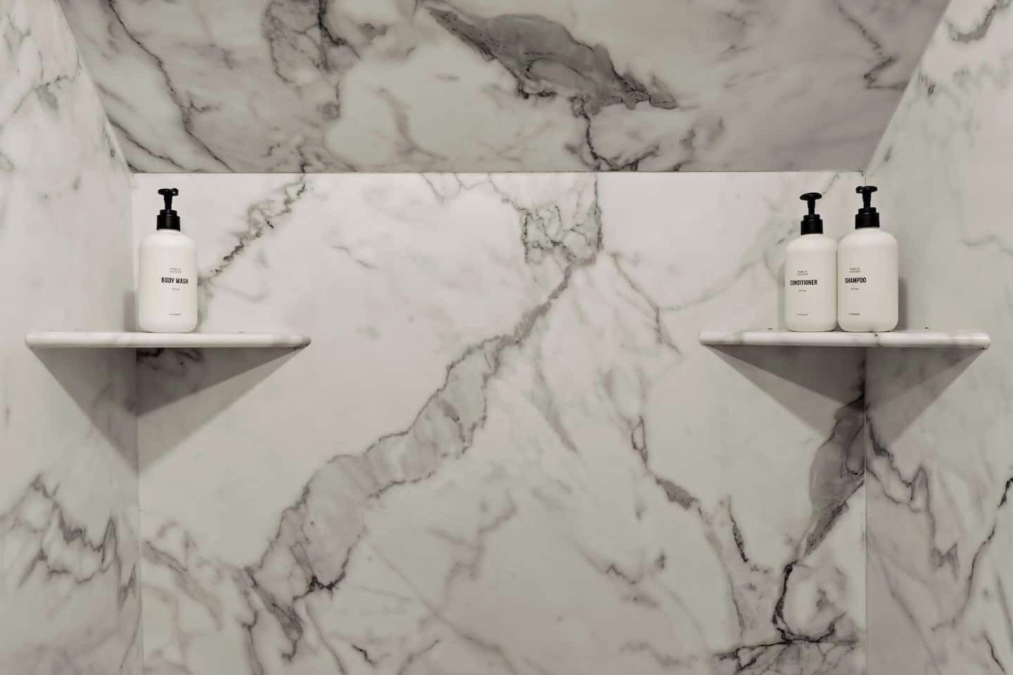 Une salle de bain aux murs recouverts de marbre, avec deux étagères blanches, chacune contenant un flacon de gel douche, d’après-shampoing et de shampoing.