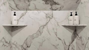 Une salle de bain aux murs recouverts de marbre, avec deux étagères blanches, chacune contenant un flacon de gel douche, d’après-shampoing et de shampoing.
