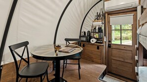 Interior acogedor de una tienda de campaña con una mesa redonda, dos sillas, un estante de madera con utensilios de cocina y una puerta que da al exterior.