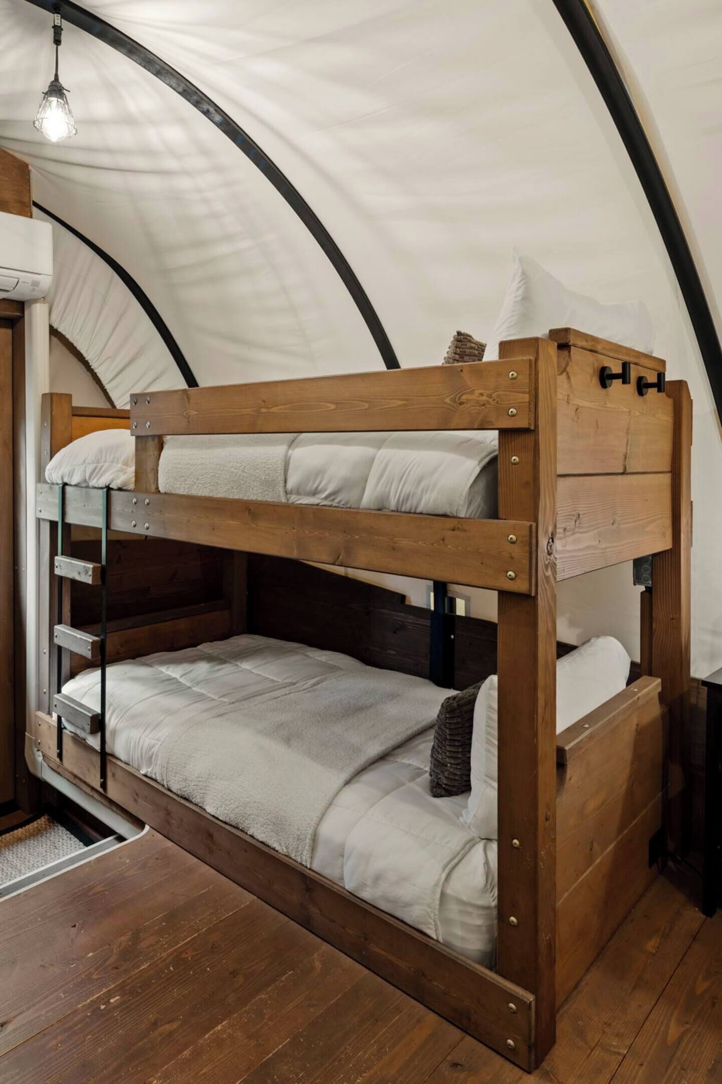 Un lit superposé avec une structure en bois et une literie blanche, installé dans une pièce ressemblant à une tente, dotée d’un plafond incurvé et d’un luminaire suspendu.
