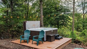 A hot tub with two chairs on a wooden deck surrounded by trees, with string lights overhead.