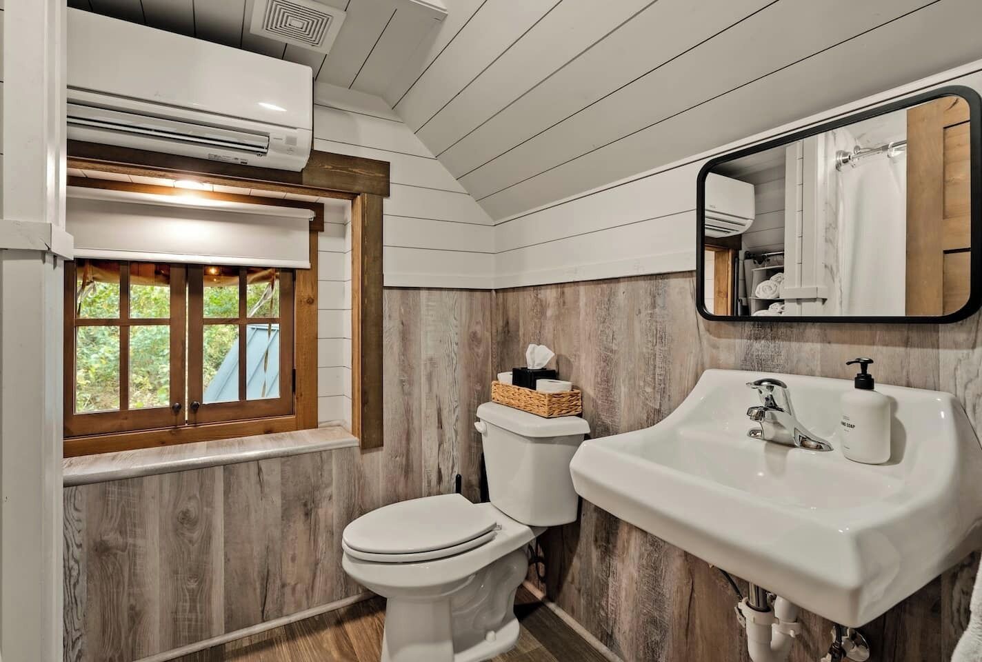 Un baño con pared de madera, ventana con persianas, inodoro, lavabo y espejo.