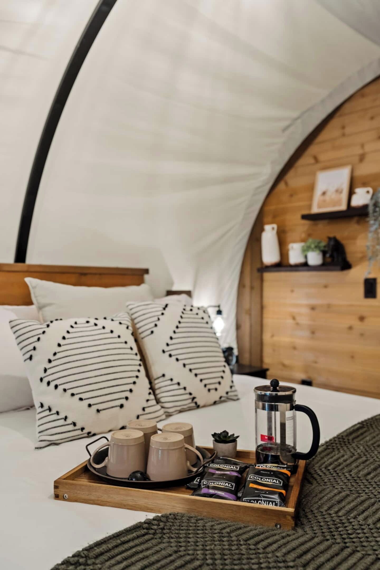 A cozy bedroom with a wooden headboard, patterned pillows, a tray with coffee mugs and tea bags, and a coffee pot.
