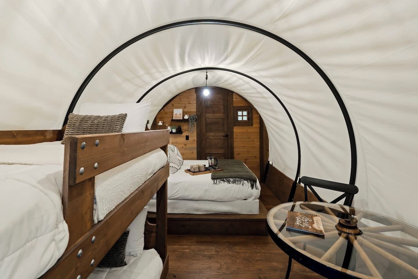 Une pièce accueillante aménagée comme une tente, avec deux lits superposés en bois, une petite table sur laquelle est posé un livre, et une porte en bois donnant sur une autre pièce.