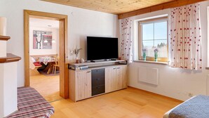 Habitació amb sostre de fusta, televisió de pantalla plana sobre un moble de fusta, finestra amb cortines florals i vista a l'exterior.