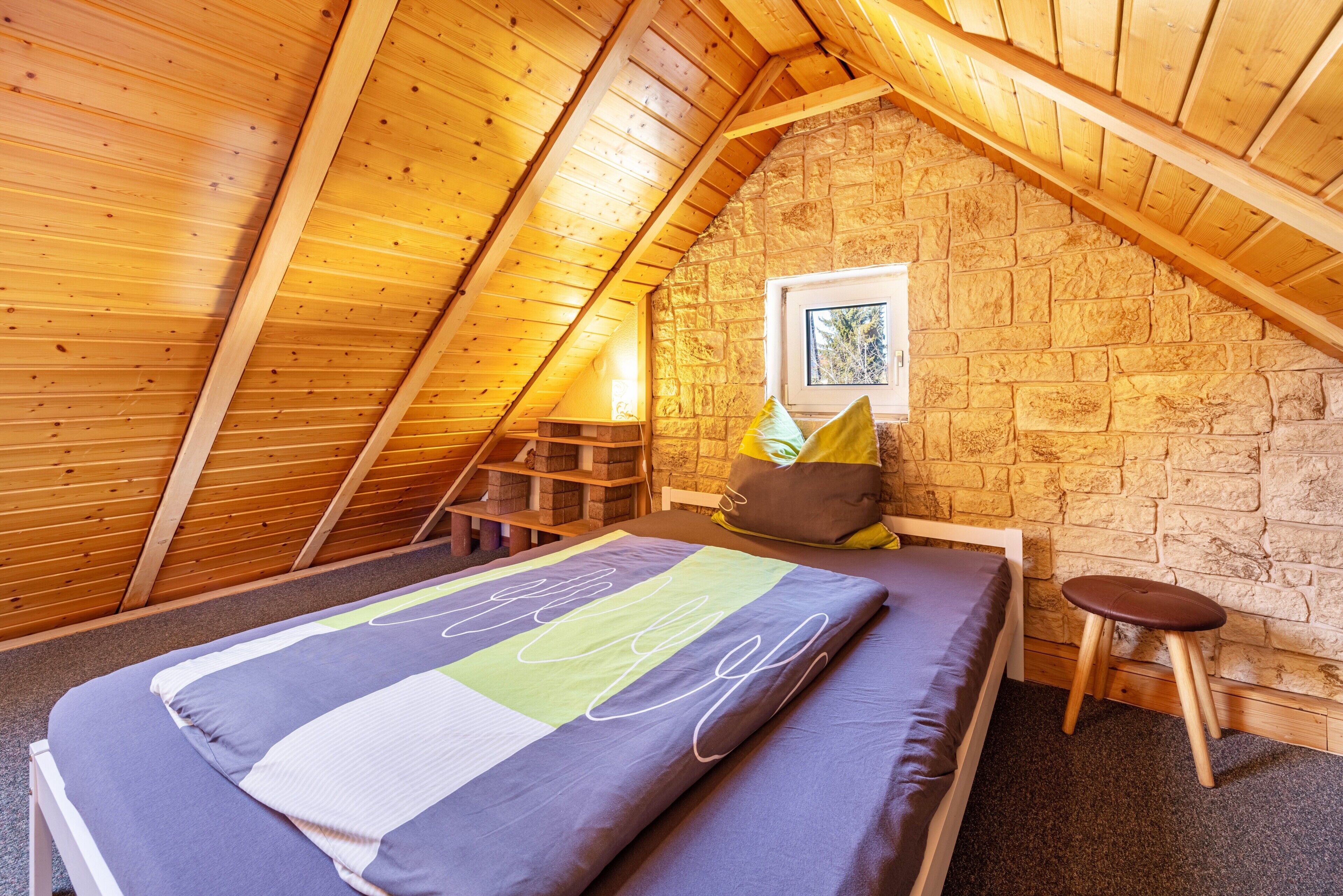 Une chambre mansardée chaleureuse avec un plafond en pente en bois, un lit simple, un petit tabouret et un mur en pierre.