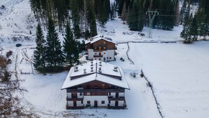 A two-story wooden chalet with a snow-covered roof, surrounded by a forested area. - El Mugo-Boscoverde 1 (Rocca Pietore)