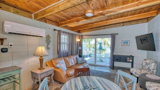 A cozy room with a wooden ceiling, a leather sofa, a round table with a crab design, a TV mounted on the wall, and a view of the outdoors through a sliding glass door. - Keys to the Sea + Cabana Club! (Key Colony Beach)