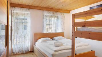 Een houten stapelbed met witte beddengoed, een houten hoofdbord en dunne gordijnen met bladmotieven.