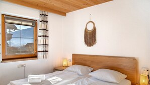 A bedroom with a wooden headboard, a bed with white linens, a window with curtains, a wall-mounted decorative item, and bedside lamps. - Holiday Home 'Haus Hart Im Zillertal 6 People' with Mountain View, Terrace & Wi-Fi (Hart im Zillertal)