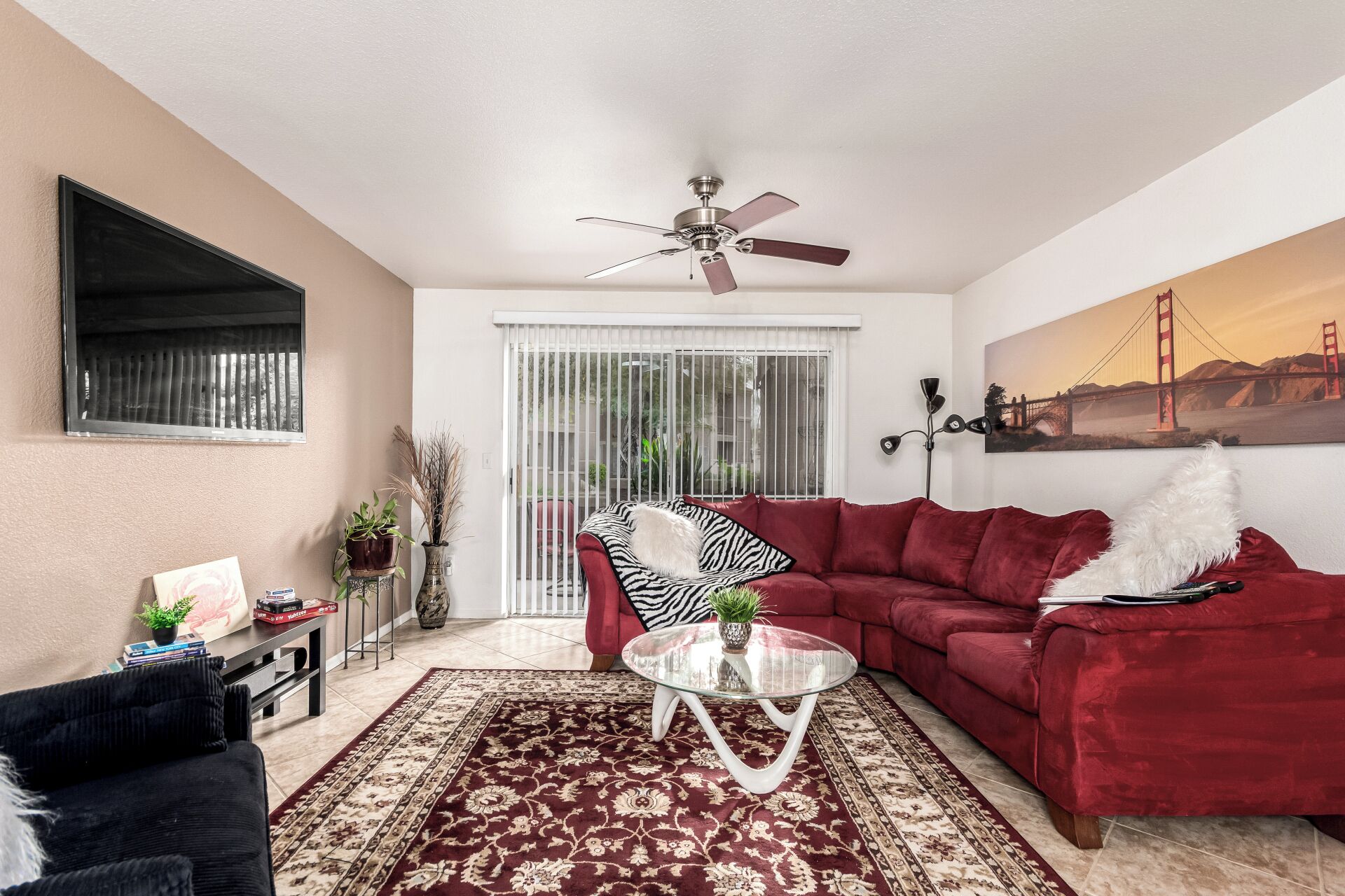 A living room with a red sofa, a glass coffee table, a black armchair, a ceiling fan, a large window with blinds, and a painting of a bridge.