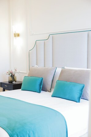 A neatly made bed with a teal bedspread and pillows, a bedside table with a vase of flowers, and a wall-mounted light fixture. - Sofia Suites &Rooms (Pompei)