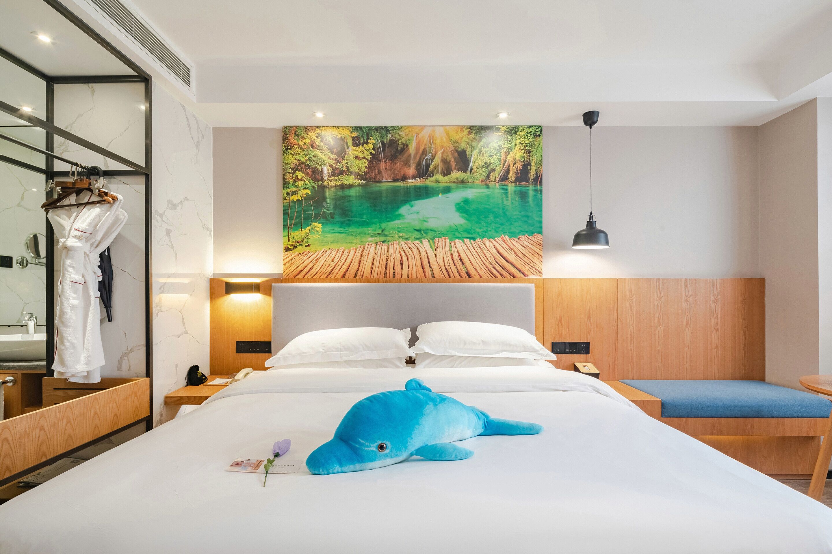 A hotel room with a large bed, a wooden headboard, a nightstand with a lamp, a wooden bench, and a large painting of a forest and lake.