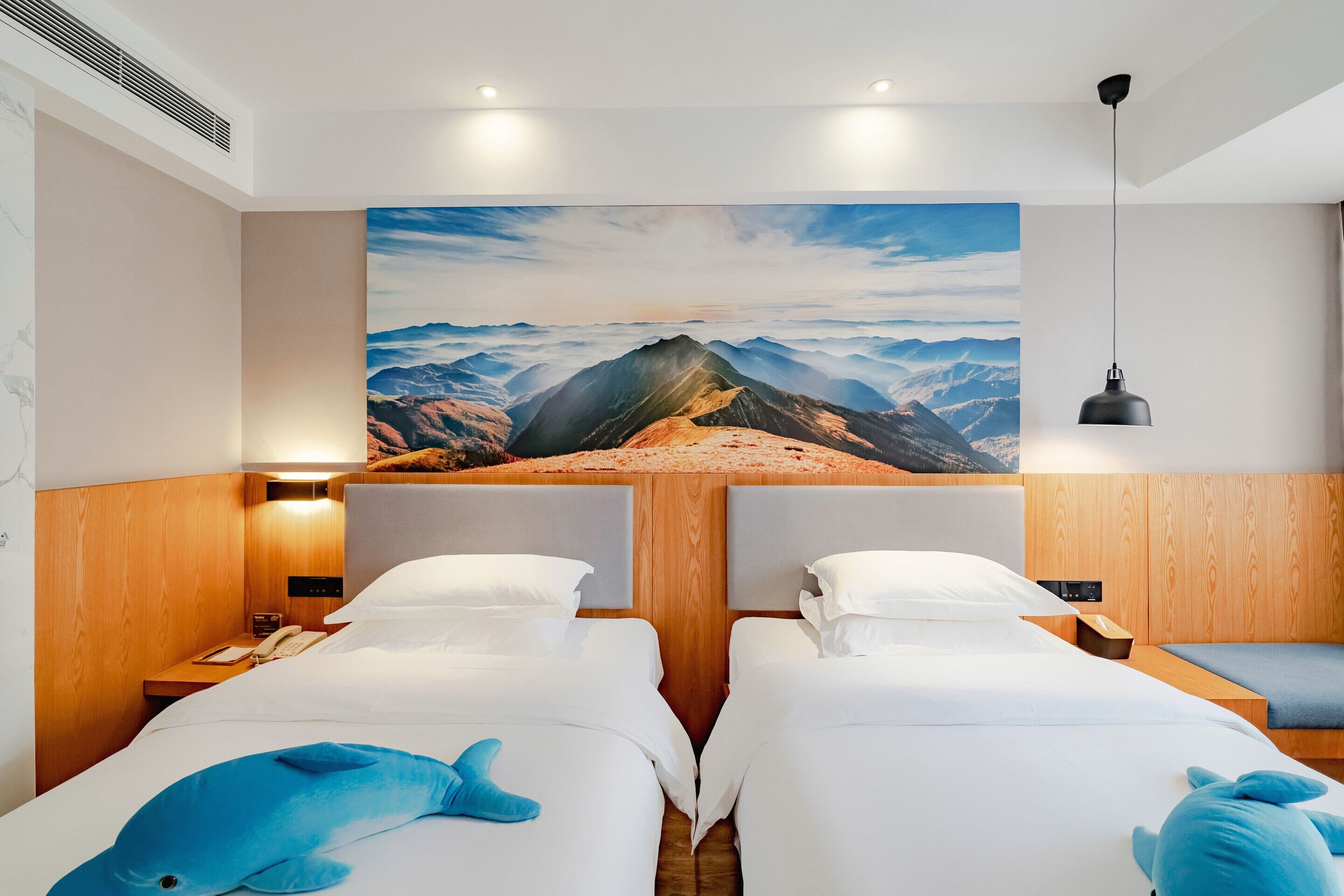 A hotel room with two beds, a large mountain view painting, wooden paneling, and a hanging pendant light.