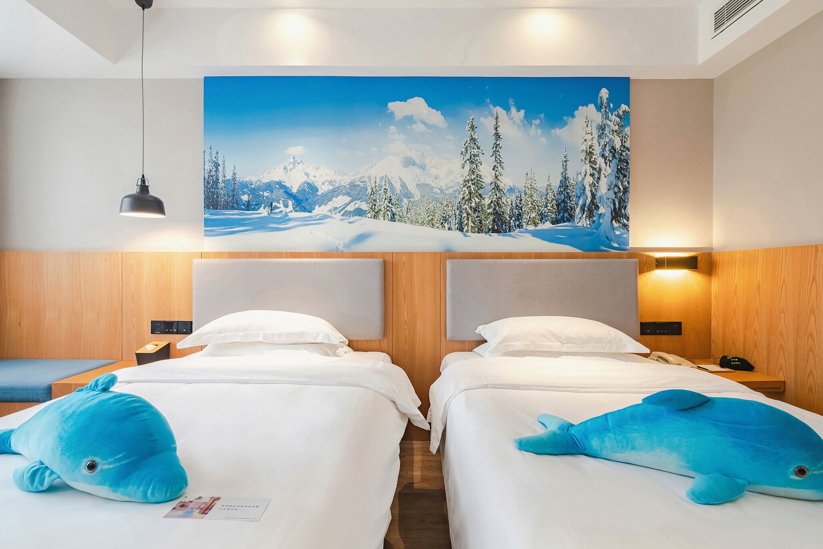 Habitación de hotel con dos camas, un cuadro grande en la pared que muestra un paisaje invernal de montañas y un peluche de ballena azul sobre cada cama.